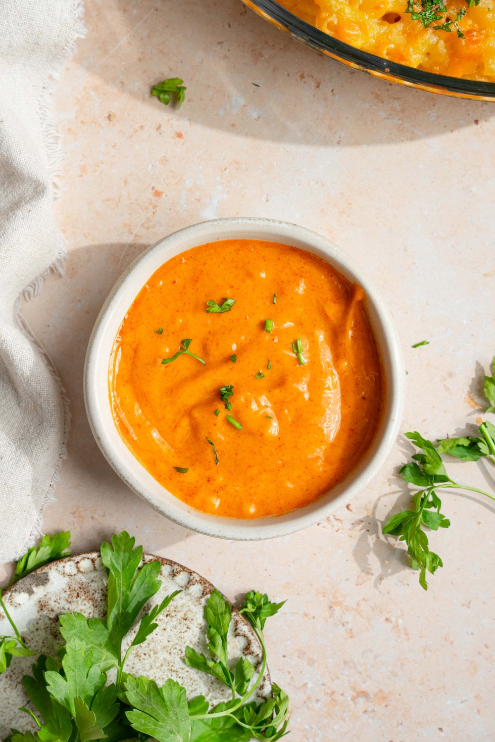 A small white bowl with campfire sauce garnished with fresh parsley. The bowl is on a tan counter with a small plate of garnishes.