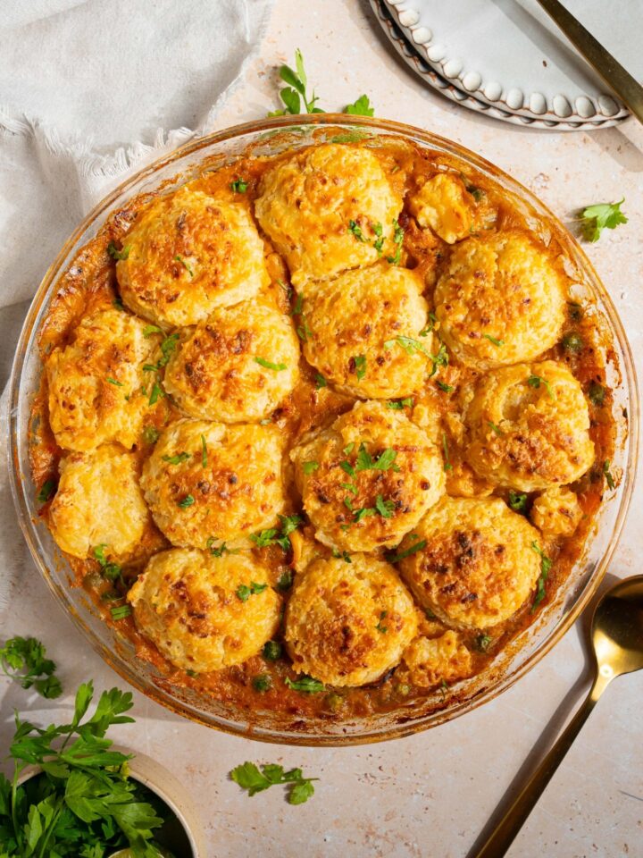 A round baking dish with baked cheddar bay biscuit chicken pot pie garnished with fresh parsley. The dish is on a tan counter with a spoon and small bowl of garnishes.