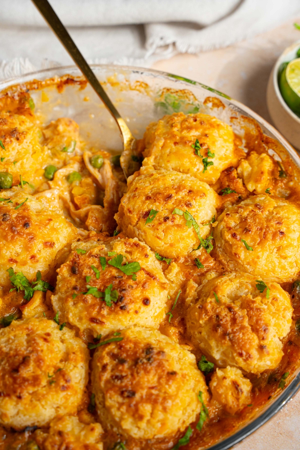 A round baking dish with baked cheddar bay biscuit chicken pot pie garnished with fresh parsley. A spoon is removing a slice from the dish. The dish is on a tan counter with a white cloth napkin.