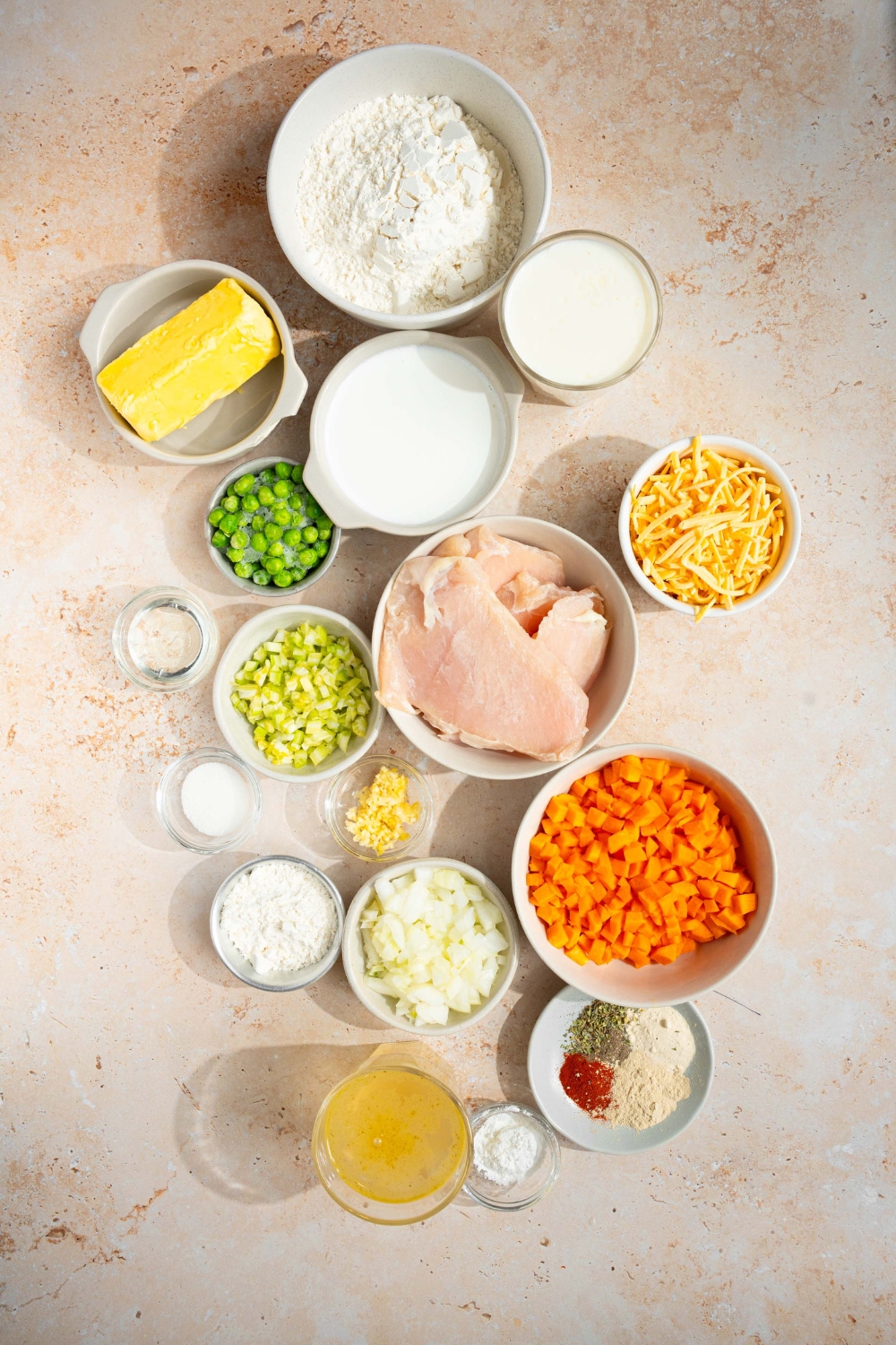 An overhead shot of several bowls in various sizes containing ingredients to make cheddar bay biscuit chicken pot pie including flour, buttermilk, shredded cheddar cheese, butter, chicken, peas, chicken broth, carrots, onion, corn, heavy cream, half and half, and seasonings.