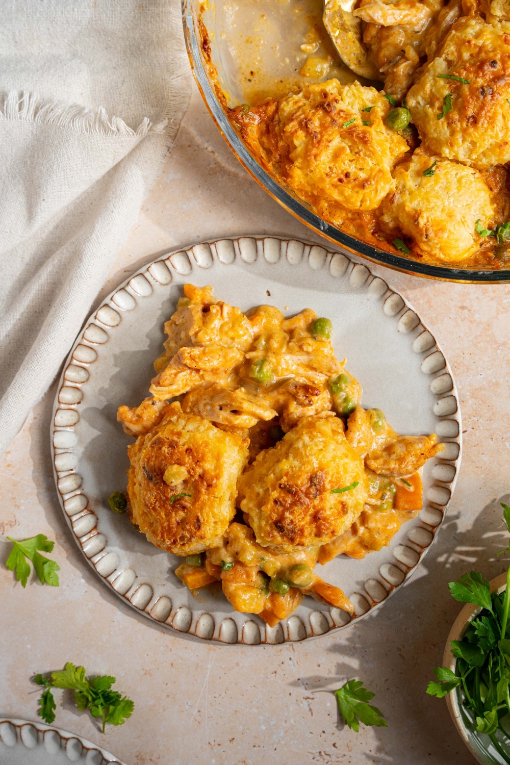 A plate with a slice of cheddar bay biscuit chicken pot pie. The plate is on a tan counter with a baking dish of pot pie, white cloth napkin, and small bowl of garnishes.