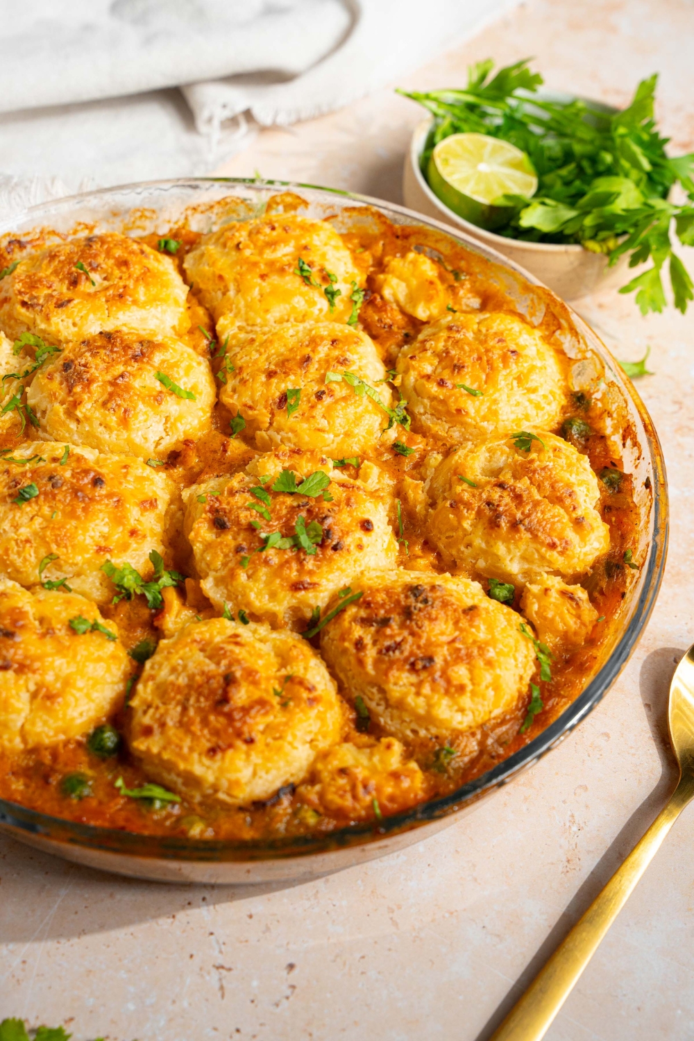 A round baking dish with baked cheddar bay biscuit chicken pot pie garnished with fresh parsley. The dish is on a tan counter with a spoon and small bowl of garnishes.