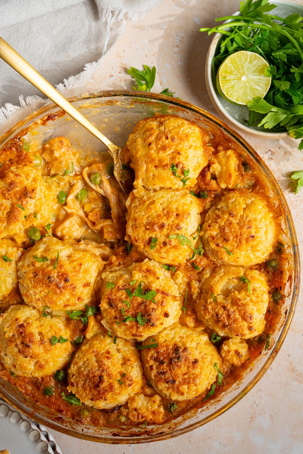 A round baking dish with baked cheddar bay biscuit chicken pot pie garnished with fresh parsley. A spoon is removing a slice from the dish. The dish is on a tan counter with a small bowl of garnishes.