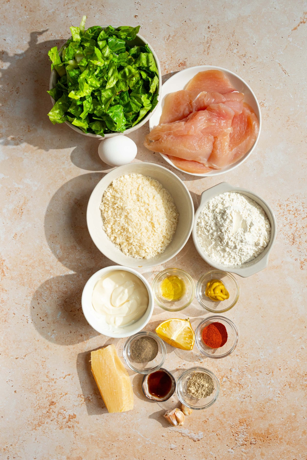 An overhead shot of several bowls in various sizes containing ingredients to make a chicken caesar sandwich including romaine lettuce, chicken breasts, parmesan cheese, flour, egg, mayo, garlic, lemon, panko bread crumbs, dijon mustard, and seasonings.