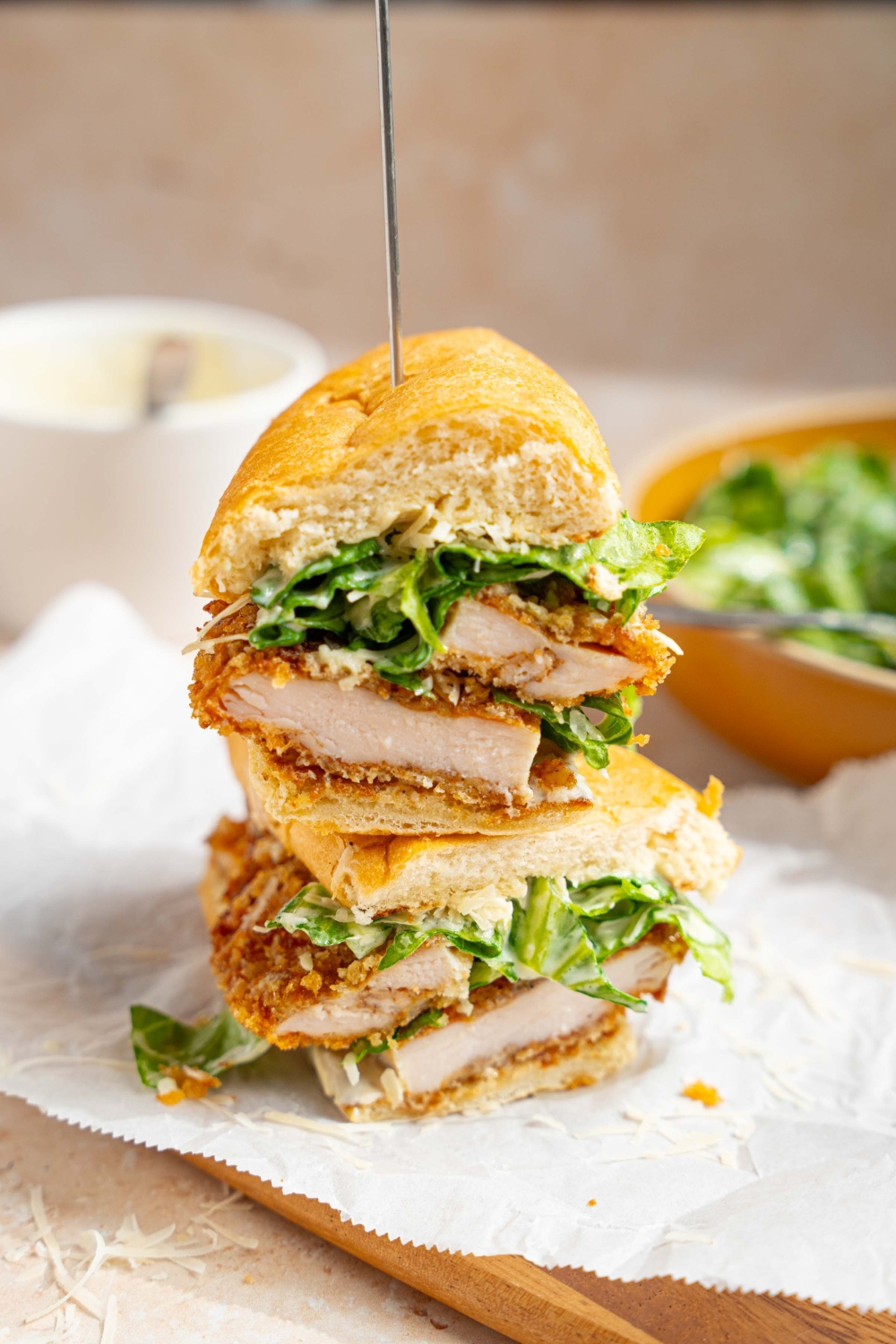 A sliced chicken caesar sandwich stacked and skewered. The sandwich is on a wooden board lined with parchment paper. The board is on a tan counter with a bowl of caesar salad.