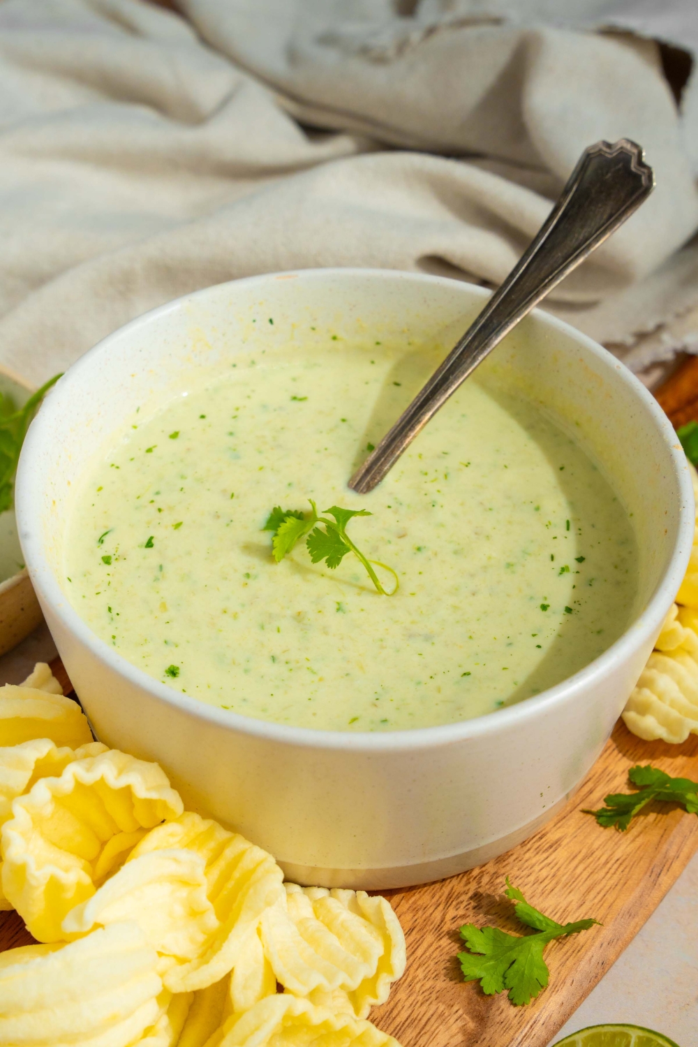 A white bowl with Chuy's jalapeno dip garnished with fresh cilantro. There is a spoon in the bowl. The bowl is on a wooden board with chips. The board is on a tan counter with a white cloth napkin.