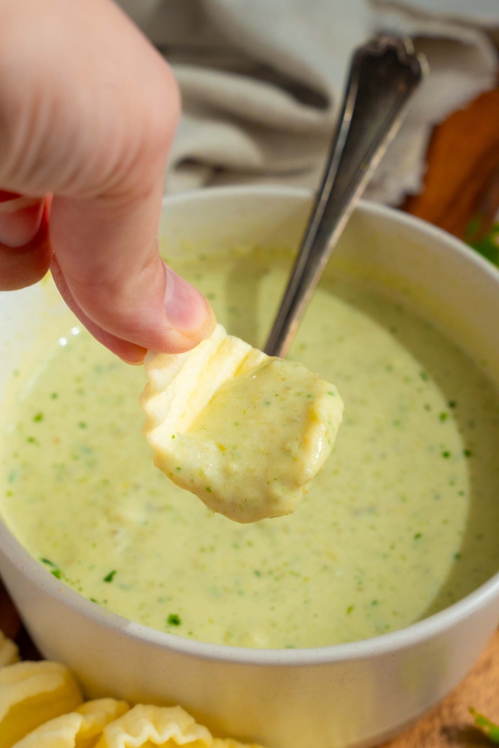 A white bowl with Chuy's jalapeno dip garnished with fresh cilantro. There is a spoon in the bowl. A hand is dipping a chip into the bowl.