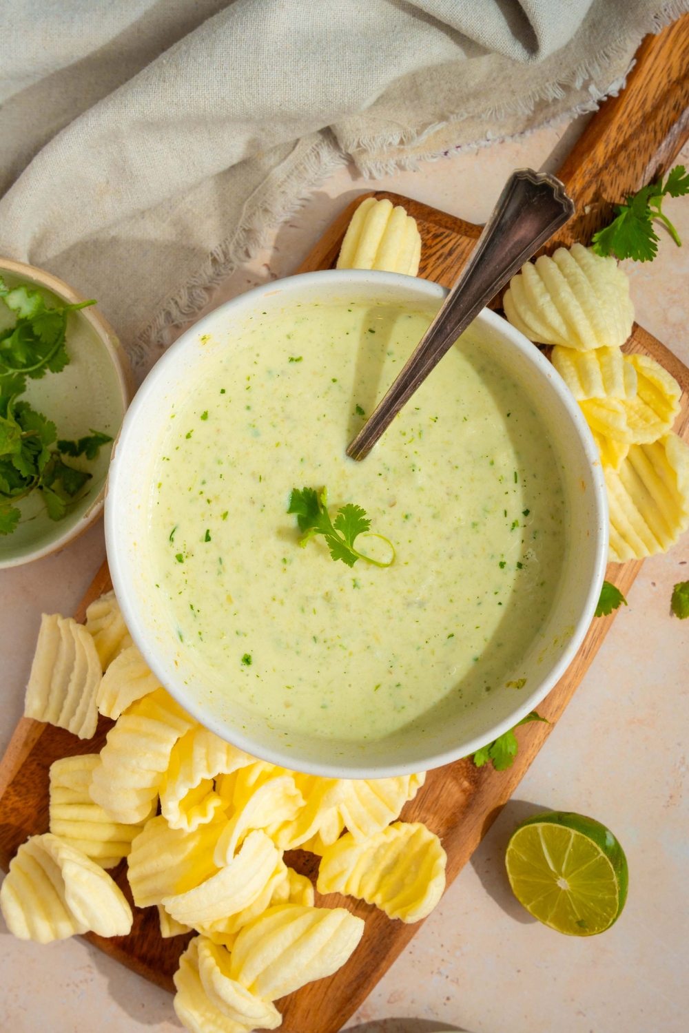 A white bowl with Chuy's jalapeno dip garnished with fresh cilantro. There is a spoon in the bowl. The bowl is on a wooden board with chips. The board is on a tan counter with a white cloth napkin and small bowl of cilantro.