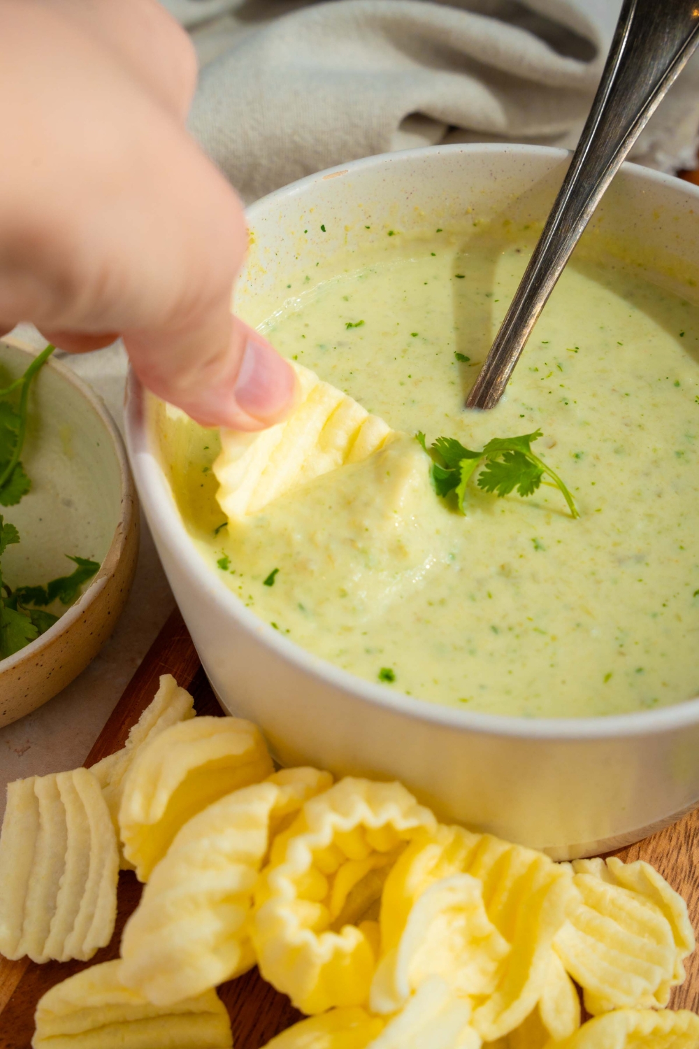 A white bowl with Chuy's jalapeno dip garnished with fresh cilantro. There is a spoon in the bowl. A hand is dipping a chip into the bowl.