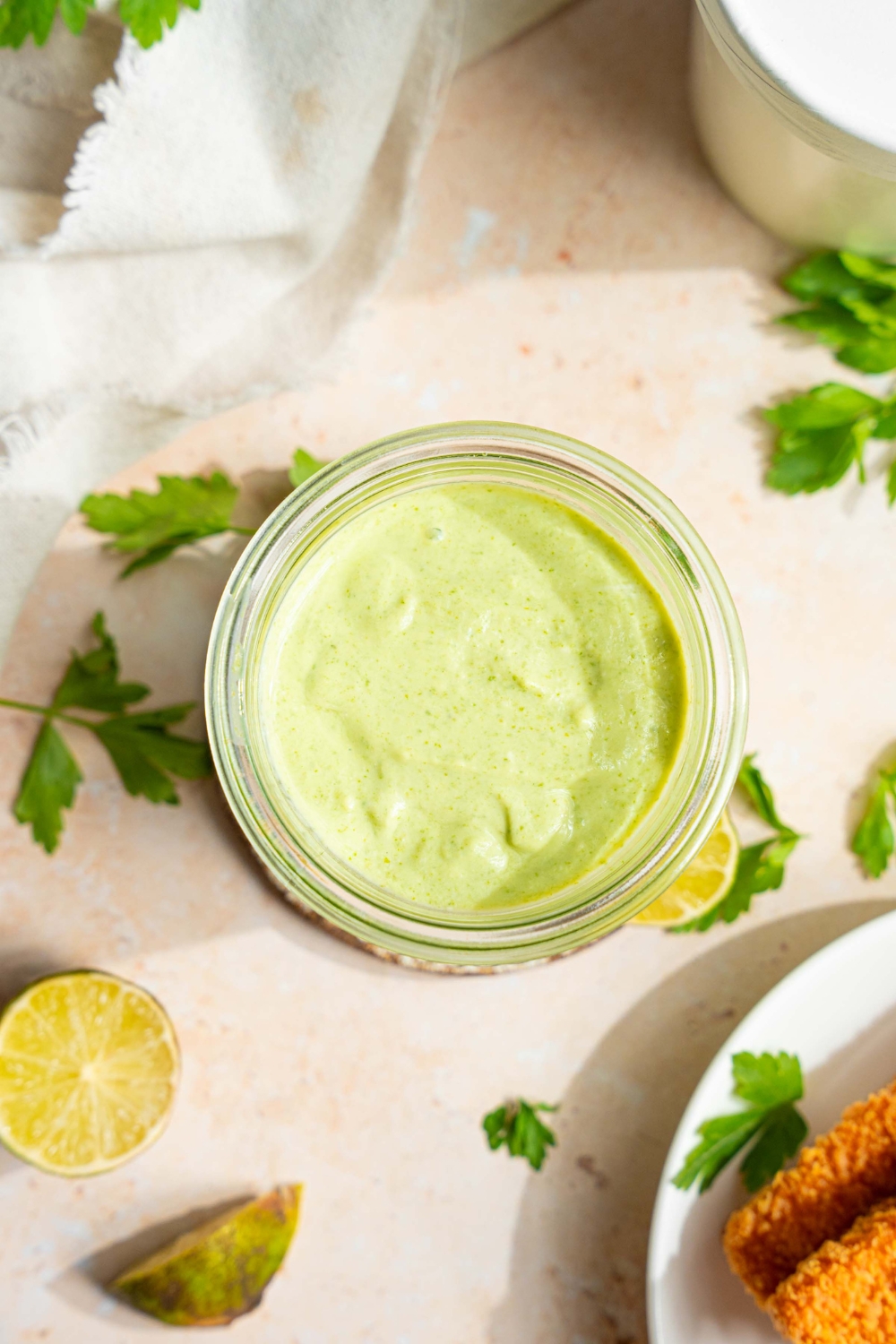 A jar of cilantro lime crema on a tan counter with sprigs of cilantro, lime wedges, and a white cloth napkin.