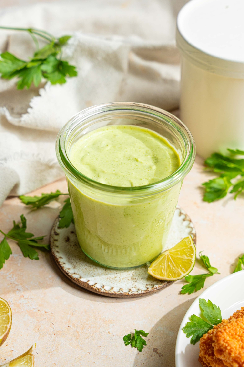 A jar of cilantro lime crema on a small plate on a tan counter with sprigs of cilantro, lime wedges, and a white cloth napkin.