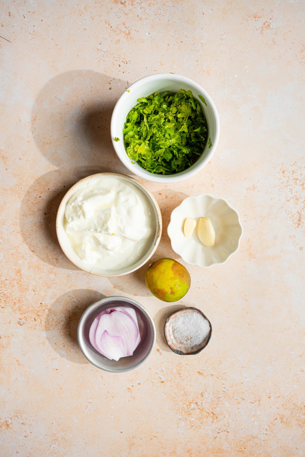 An overhead shot of several bowls in various sizes containing ingredients to make cilantro lime crema including cilantro, lime, greek yogurt, garlic, red onion, and salt.