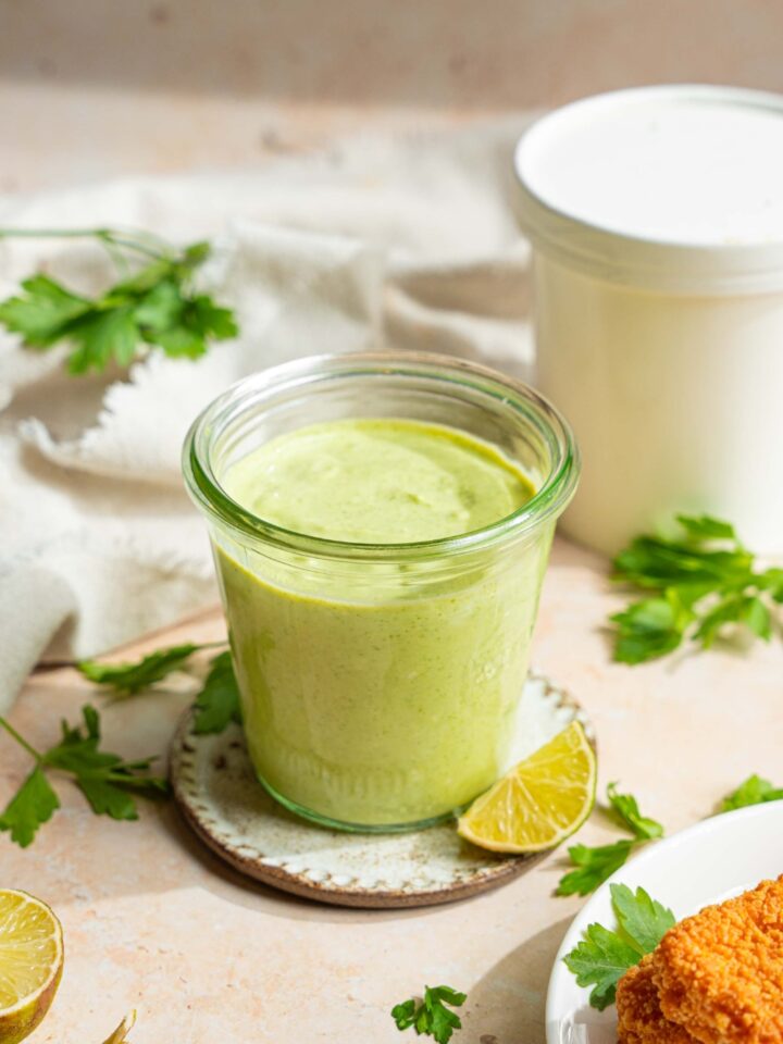 A jar of cilantro lime crema on a small plate on a tan counter with sprigs of cilantro, lime wedges, and a white cloth napkin.