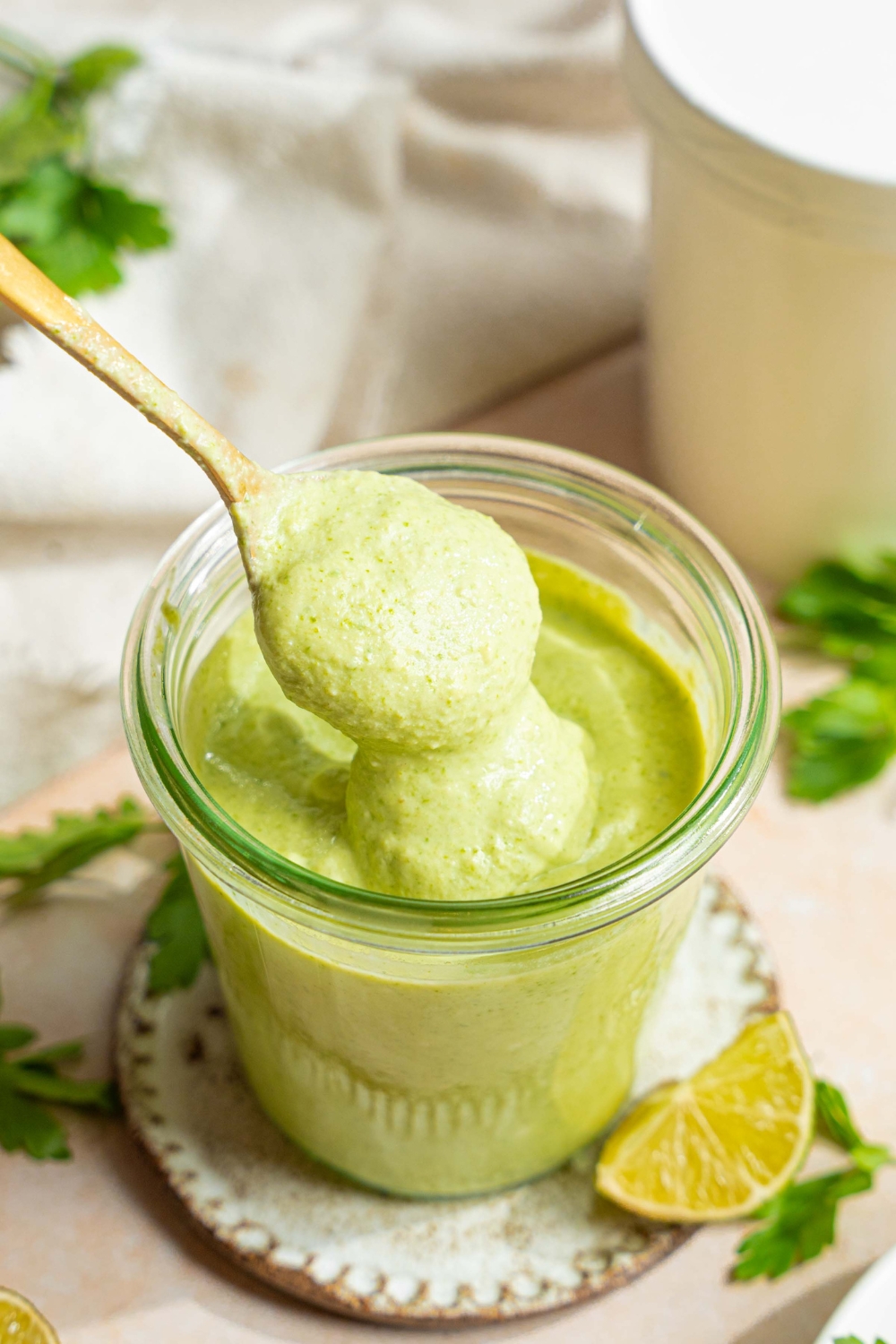 A jar of cilantro lime crema on a small plate with a spoon dipping into the jar. The plate is on a tan counter with sprigs of cilantro, lime wedges, and a white cloth napkin.