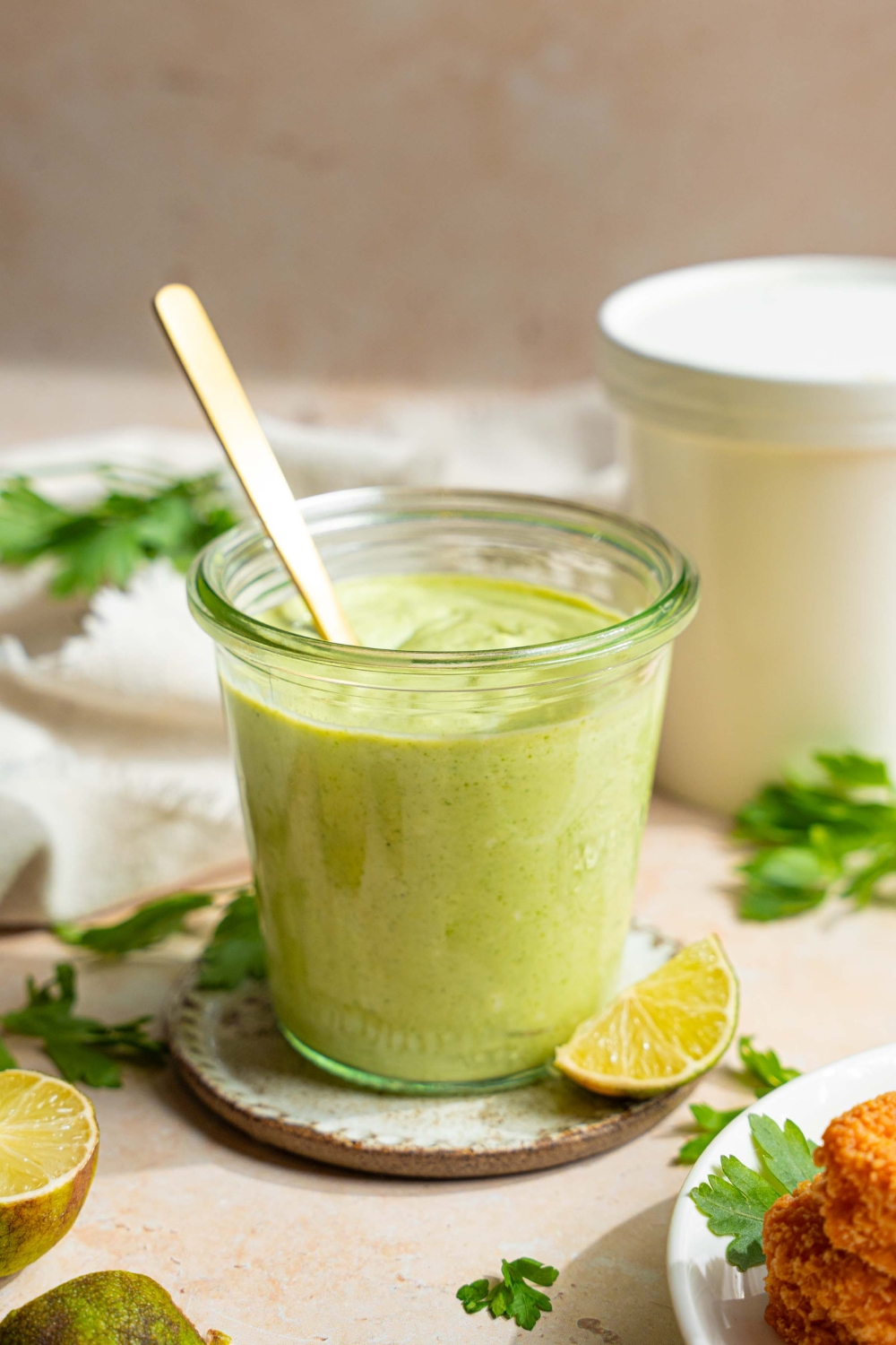 A jar of cilantro lime crema on a small plate with a spoon in the jar. The plate is on a tan counter with sprigs of cilantro, lime wedges, and a white cloth napkin.