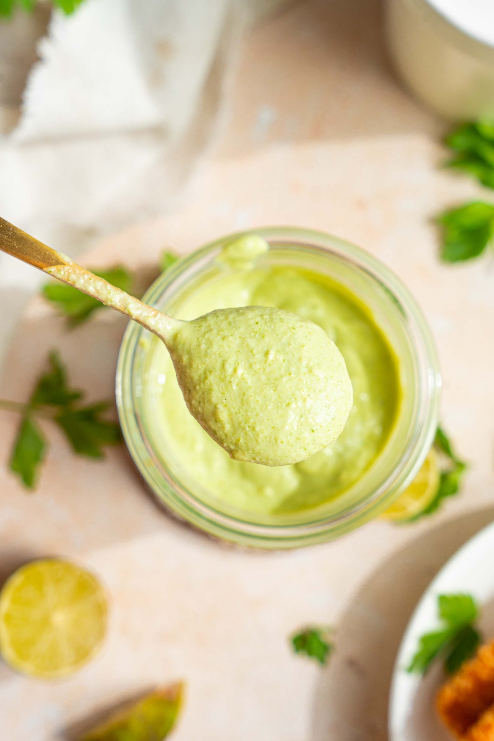 A close up of a spoon dipped in cilantro lime crema with a jar of crema on a tan counter blurred in the background.