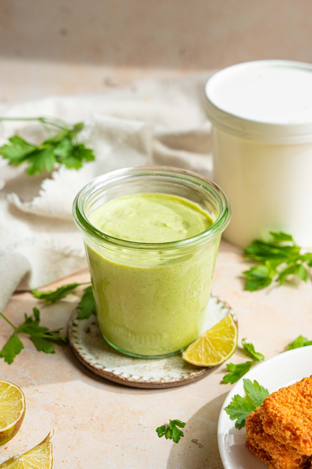 A jar of cilantro lime crema on a small plate on a tan counter with sprigs of cilantro, lime wedges, and a white cloth napkin.