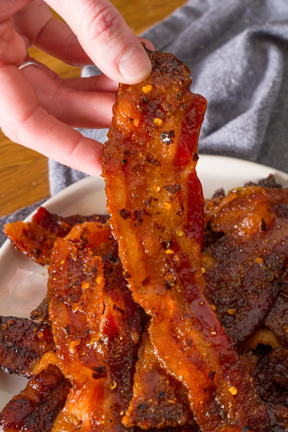 A white plate with brown sugar bacon on a wooden counter with a blue cloth napkin. A hand is taking a strip of bacon.