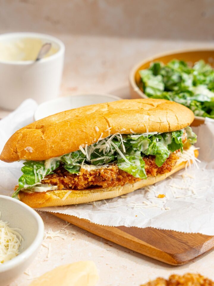 A whole chicken caesar sandwich on a wooden board lined with parchment paper. The board is on a tan counter with a bowl of caesar salad, bowl of caesar dressing, and bowl of shredded parmesan cheese.