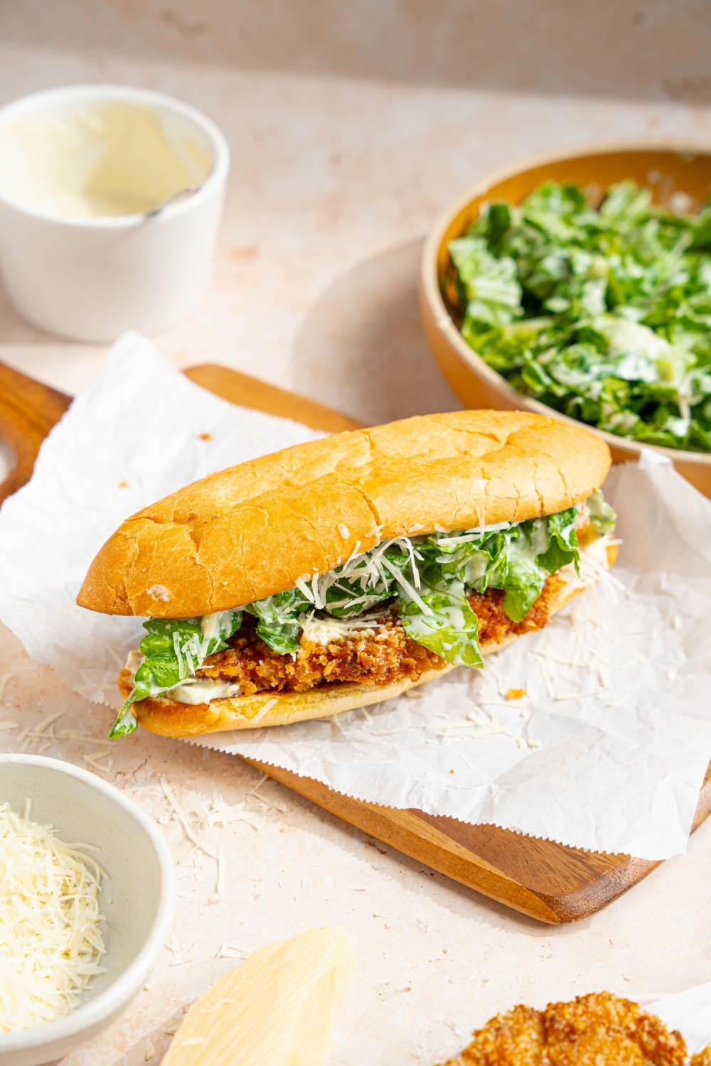 A whole chicken caesar sandwich on a wooden board lined with parchment paper. The board is on a tan counter with a bowl of caesar salad, bowl of caesar dressing, and bowl of shredded parmesan cheese.