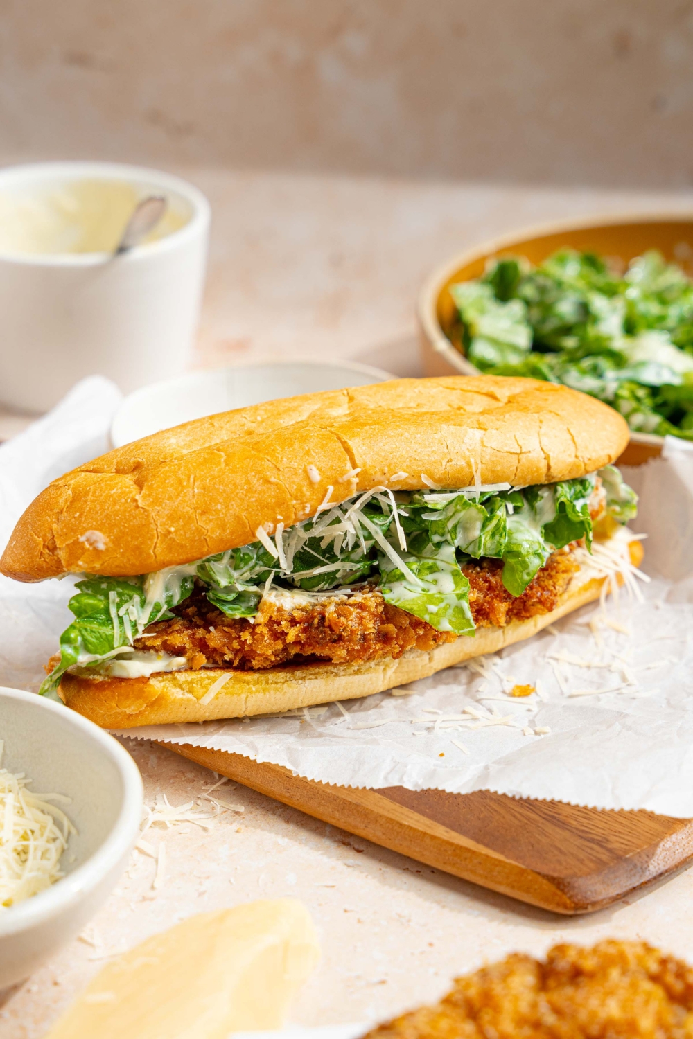 A whole chicken caesar sandwich on a wooden board lined with parchment paper. The board is on a tan counter with a bowl of caesar salad, bowl of caesar dressing, and bowl of shredded parmesan cheese.