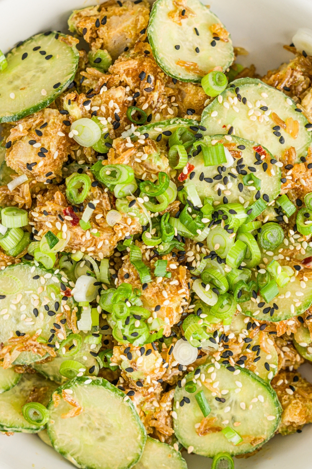 A white bowl with crispy rice chicken and cucumber salad garnished with sliced green onion and sesame seeds. There is a fork with a bite of salad over the bowl.