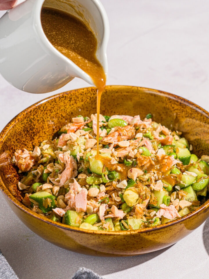 A bowl of crispy rice salmon salad with a hand pouring a saucer of creamy honey ginger dressing over the salad. The bowl is on a marble counter.