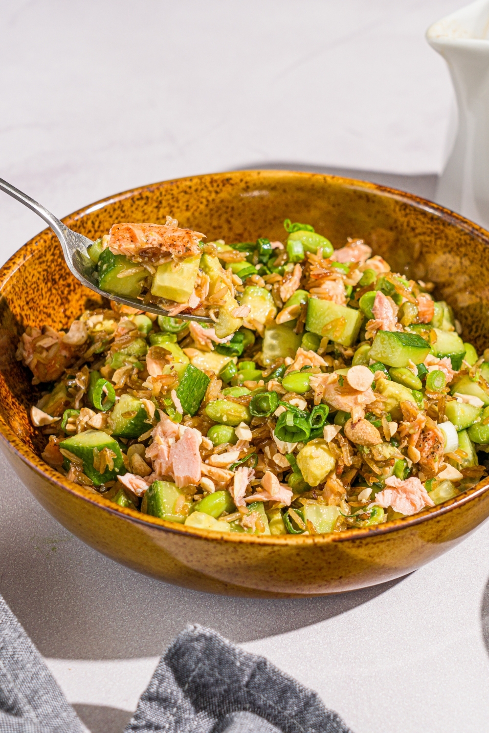 A bowl of crispy rice salmon salad with honey ginger dressing garnished with sliced green onion. There is a fork with a bite of salad over the bowl. The bowl is on a marble counter with a gray cloth napkin.