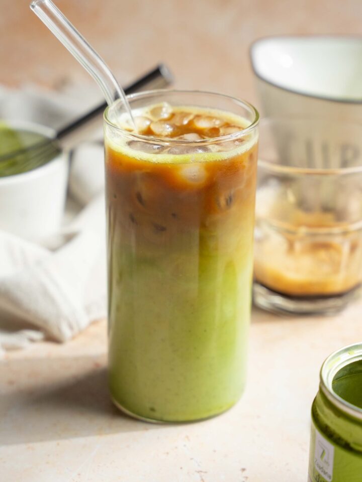 A tall glass of iced matcha latte with a glass straw. The glass is on a tan counter with a tin of matcha powder, small glass of poured espresso, and a small bowl and whisk with matcha.
