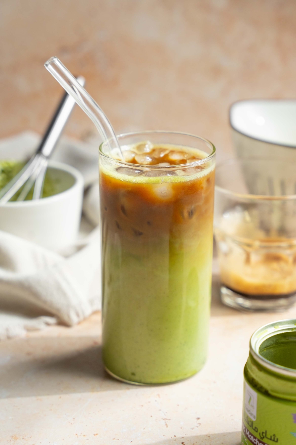 A tall glass of iced matcha latte with a glass straw. The glass is on a tan counter with a tin of matcha powder, small glass of poured espresso, and a small bowl and whisk with matcha.