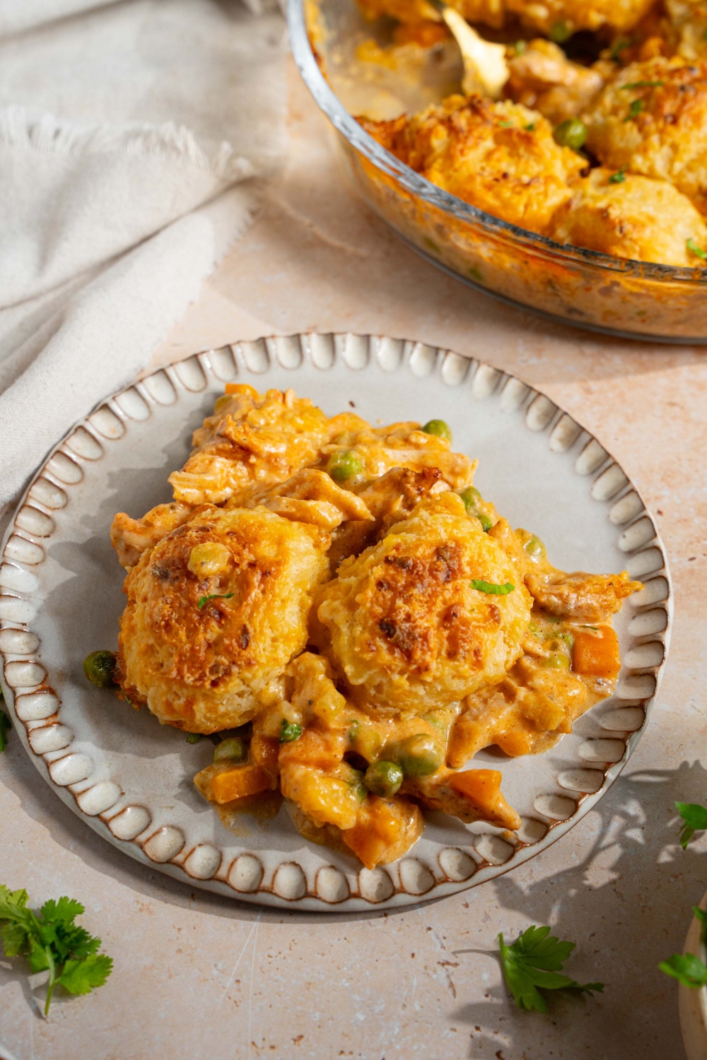 A plate with a slice of cheddar bay biscuit chicken pot pie. The plate is on a tan counter with a baking dish of pot pie and white cloth napkin.