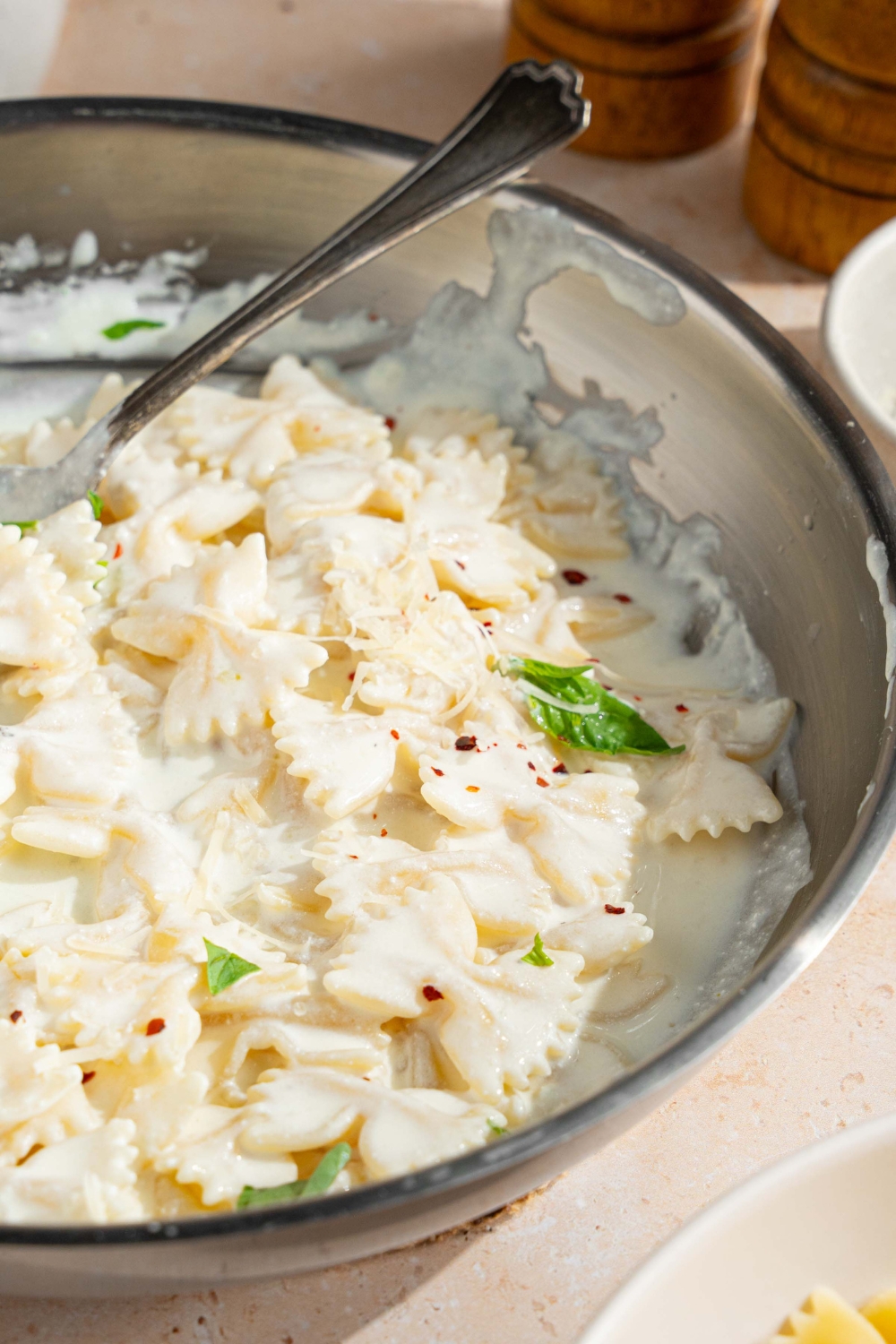 A skillet with bowtie pasta tossed in alfredo sauce. The pasta is garnished with fresh parsley and crushed red pepper. There is a fork in the pasta. The skillet is on a tan counter with a salt and pepper mill.