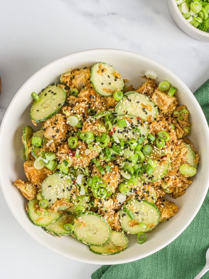 A white bowl with crispy rice chicken and cucumber salad garnished with sliced green onion and sesame seeds. The bowl is on a marble counter with small bowls of garnishes and a green cloth napkin.