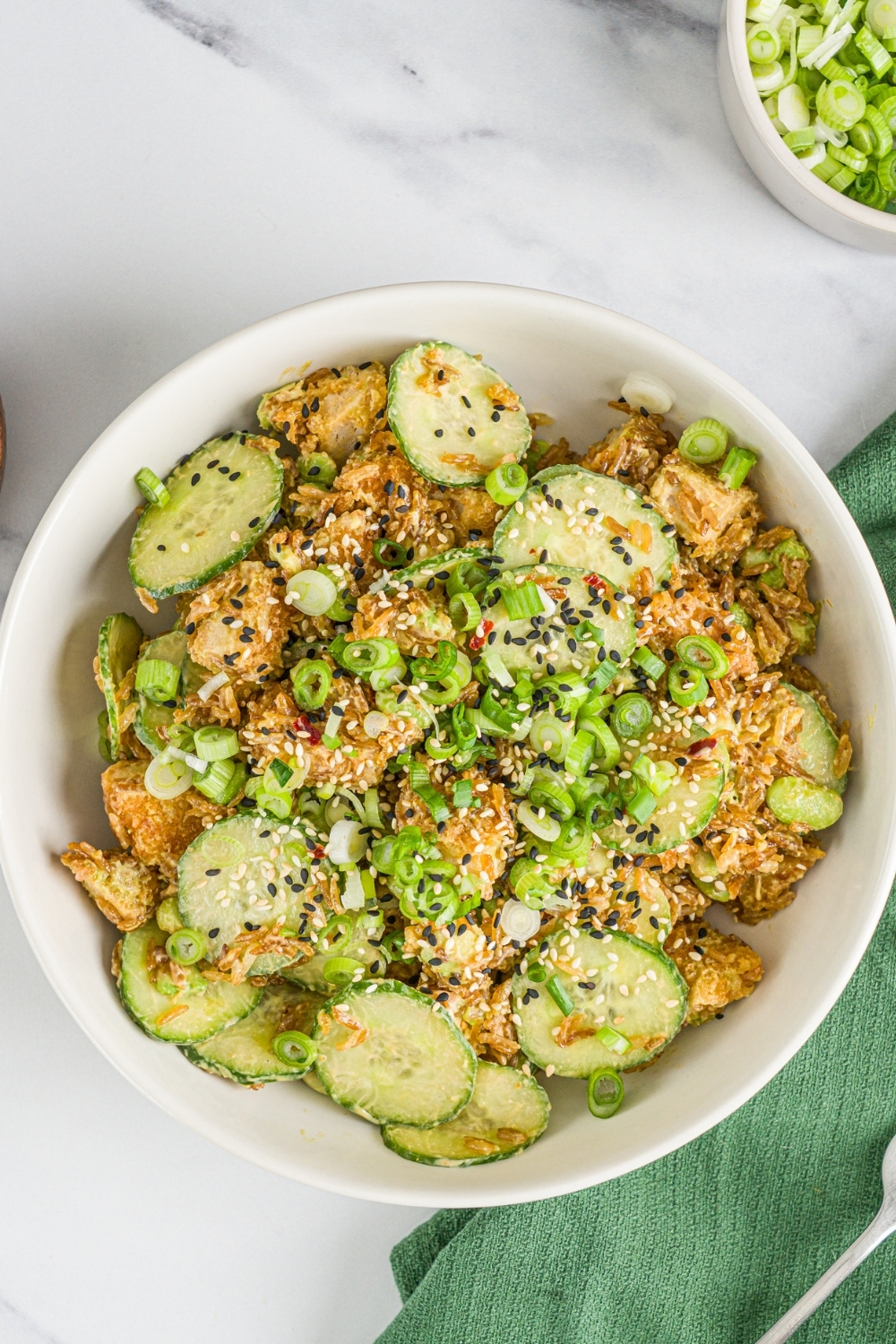 A white bowl with crispy rice chicken and cucumber salad garnished with sliced green onion and sesame seeds. The bowl is on a marble counter with small bowls of garnishes and a green cloth napkin.