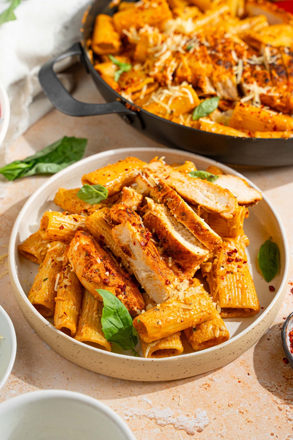 A white plate with garlic parmesan chicken pasta garnished with fresh basil and shredded parmesan and red pepper flakes. The plate is on a tan counter with a skillet of pasta and small plate of garnishes.