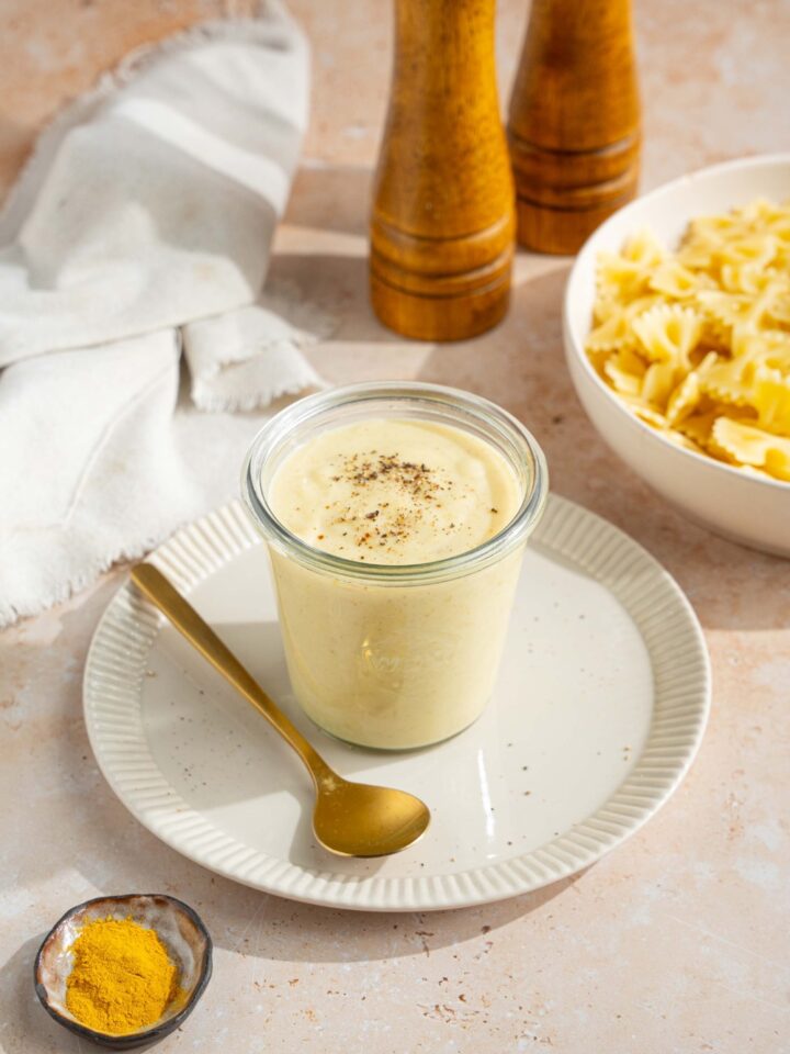 A white plate with a jar of hello fresh cream sauce garnished with a spice blend. There is a spoon on the plate. The plate is on a tan counter with a white cloth napkin and bowl of bowtie pasta.