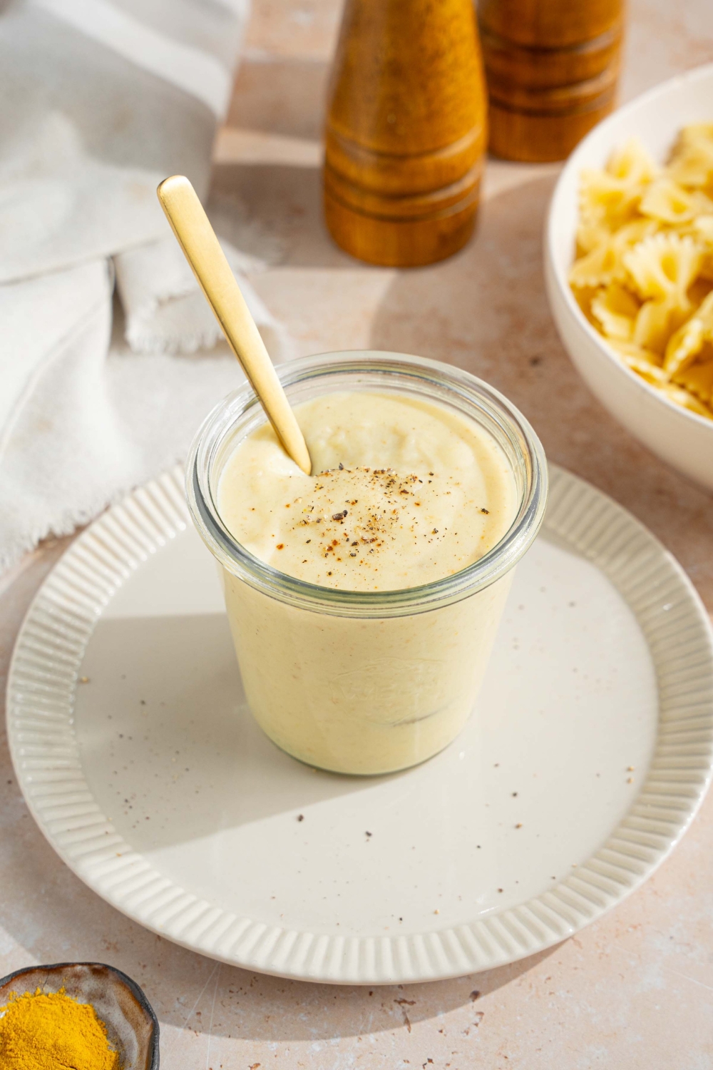 A white plate with a jar of hello fresh cream sauce garnished with a spice blend. There is a spoon in the jar. The plate is on a tan counter with a white cloth napkin and bowl of bowtie pasta.