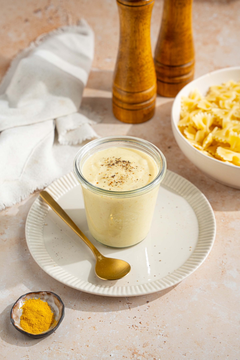 A white plate with a jar of hello fresh cream sauce garnished with a spice blend. There is a spoon on the plate. The plate is on a tan counter with a white cloth napkin and bowl of bowtie pasta.