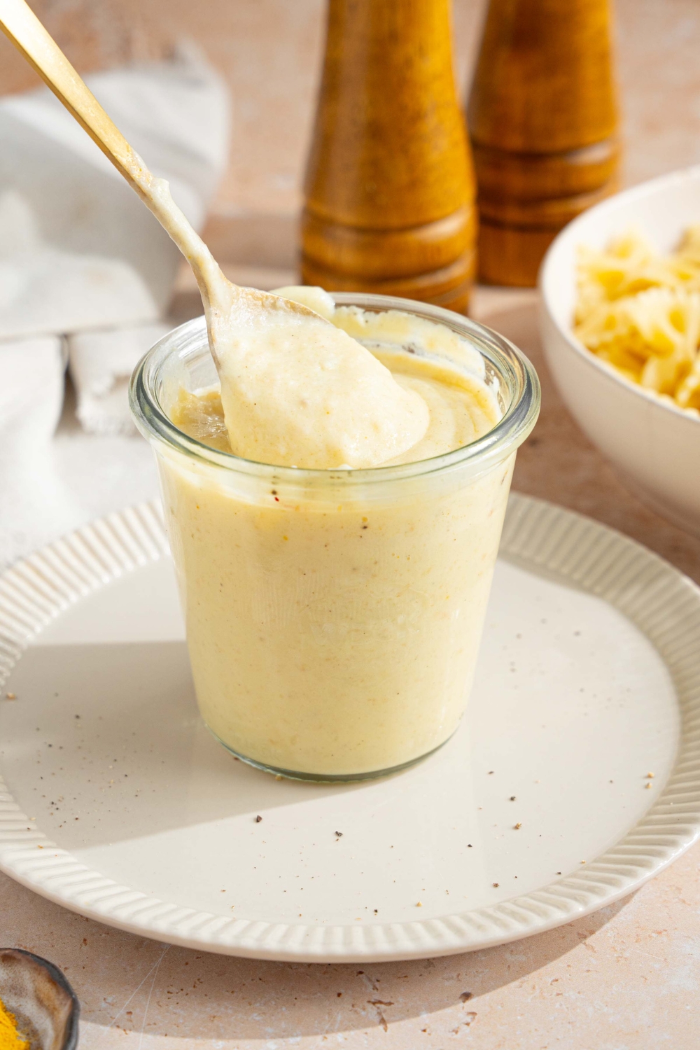 A white plate with a jar of hello fresh cream sauce garnished with a spice blend. A spoon is stirring the jar. The plate is on a tan counter with a white cloth napkin and bowl of bowtie pasta.