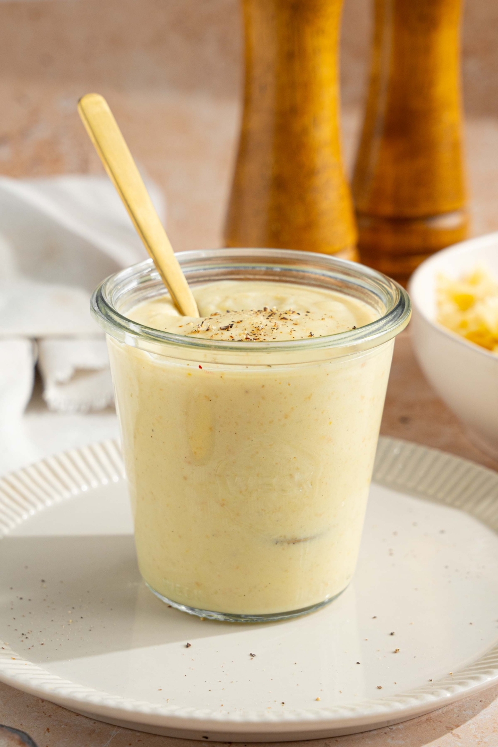 A white plate with a jar of hello fresh cream sauce garnished with a spice blend. There is a spoon in the jar. The plate is on a tan counter with a white cloth napkin and bowl of bowtie pasta.