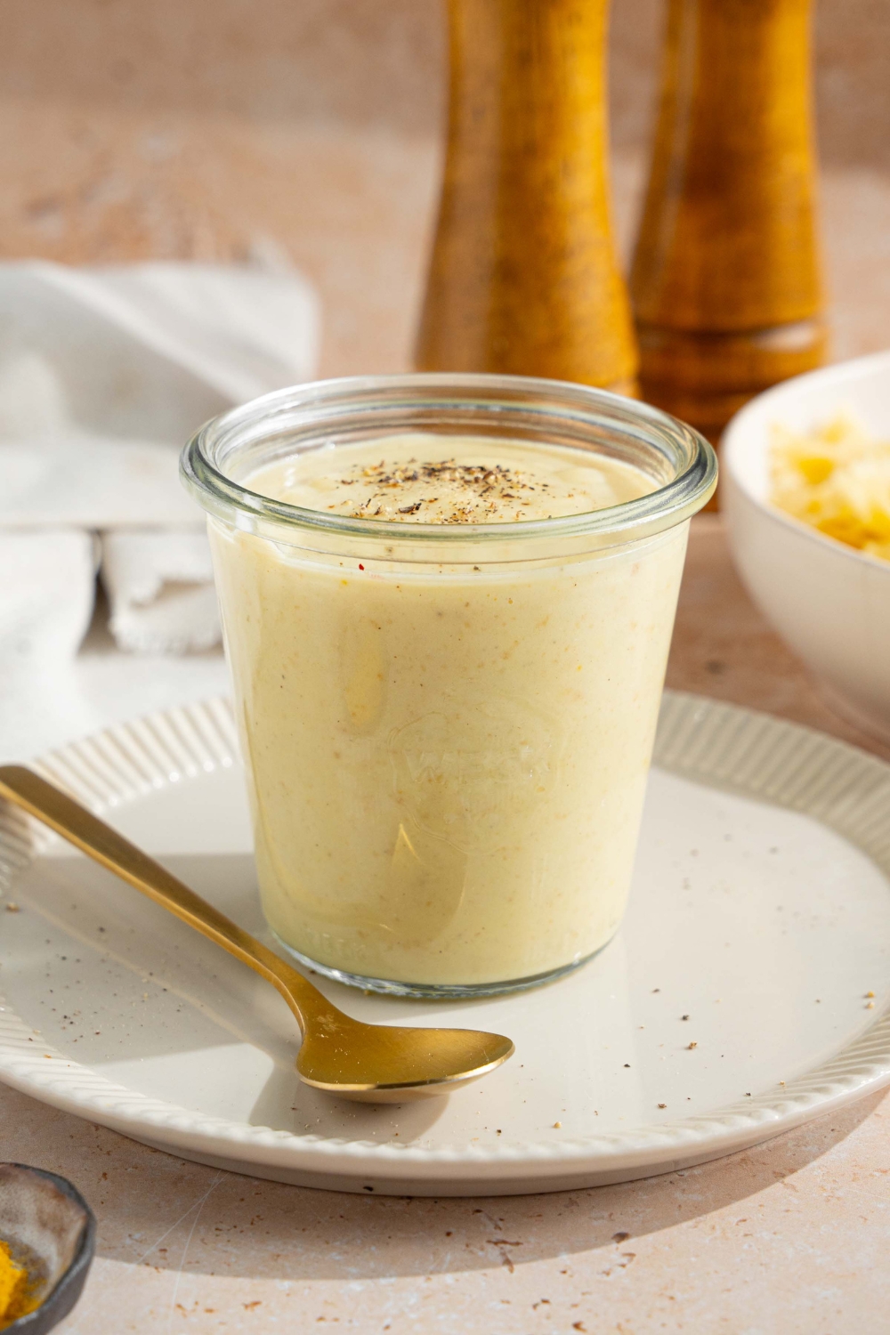 A white plate with a jar of hello fresh cream sauce garnished with a spice blend. A spoon is on the plate. The plate is on a tan counter with a white cloth napkin and bowl of bowtie pasta.