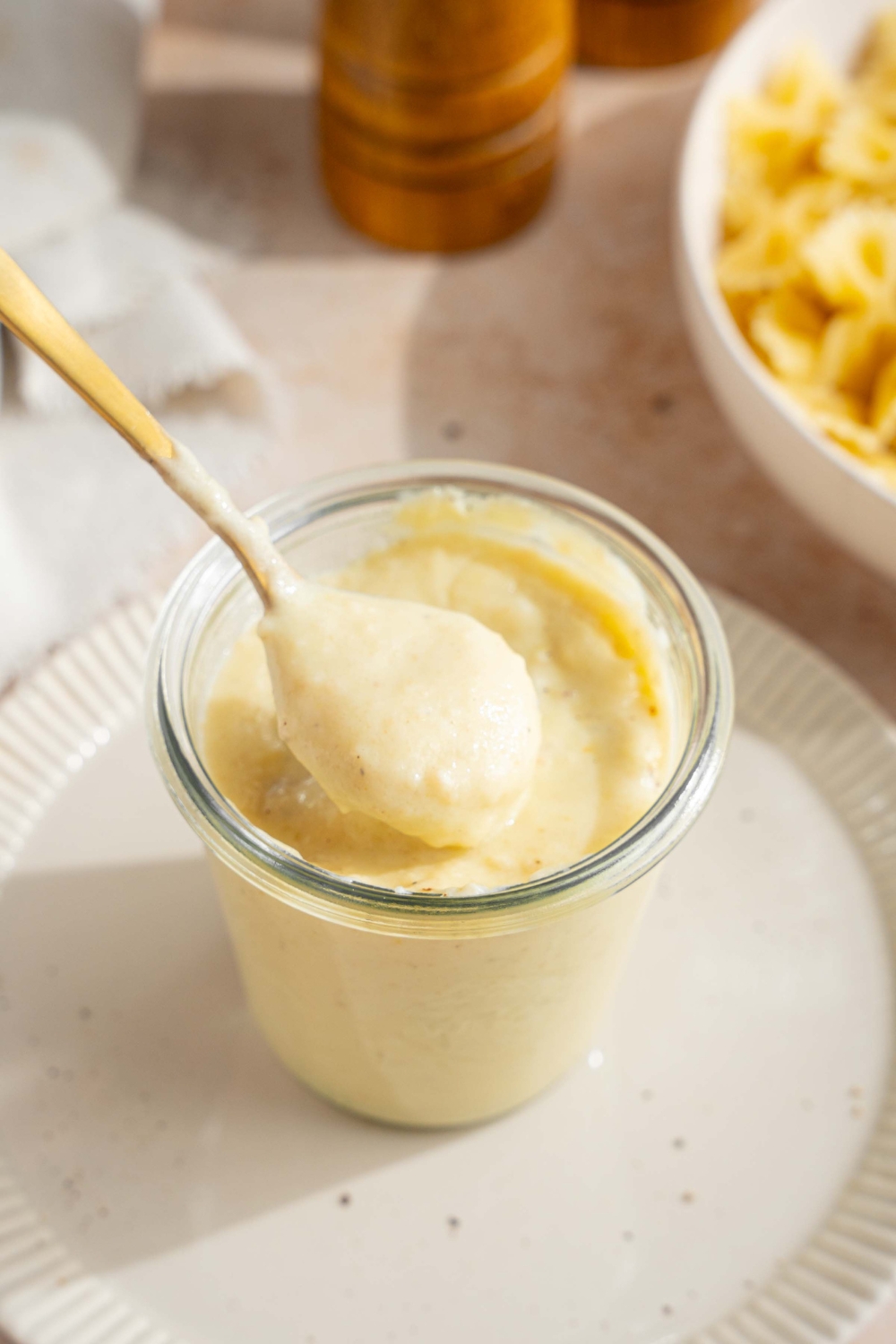 A white plate with a jar of hello fresh cream sauce garnished with a spice blend. A spoon is stirring the jar. The plate is on a tan counter with a white cloth napkin and bowl of bowtie pasta.