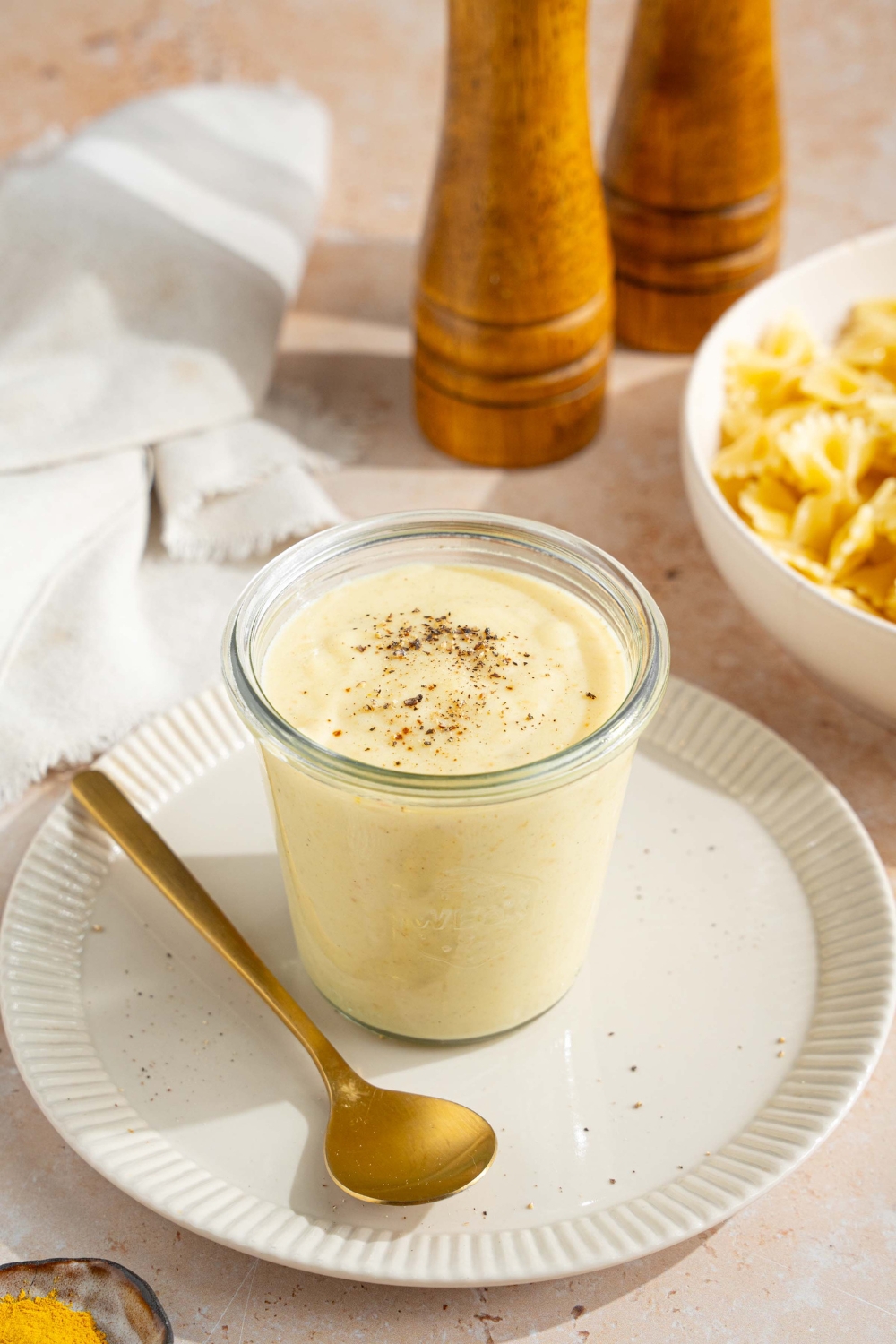 A white plate with a jar of hello fresh cream sauce garnished with a spice blend. A spoon is on the plate. The plate is on a tan counter with a white cloth napkin and bowl of bowtie pasta.