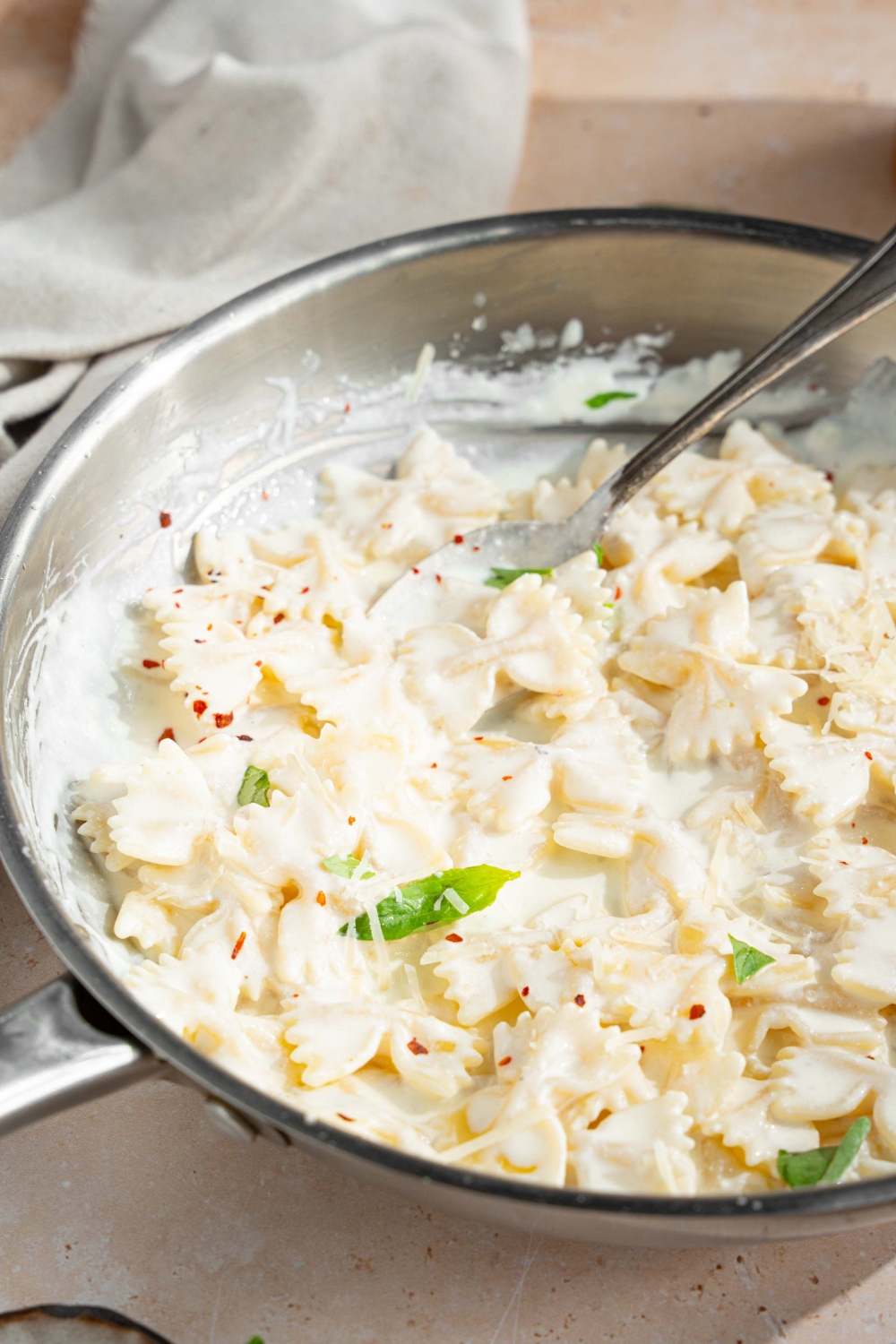 A skillet with bowtie pasta tossed in alfredo sauce. The pasta is garnished with fresh parsley and crushed red pepper. There is a fork in the pasta. The skillet is on a tan counter with a bowl of garlic and bowl of bowtie pasta.