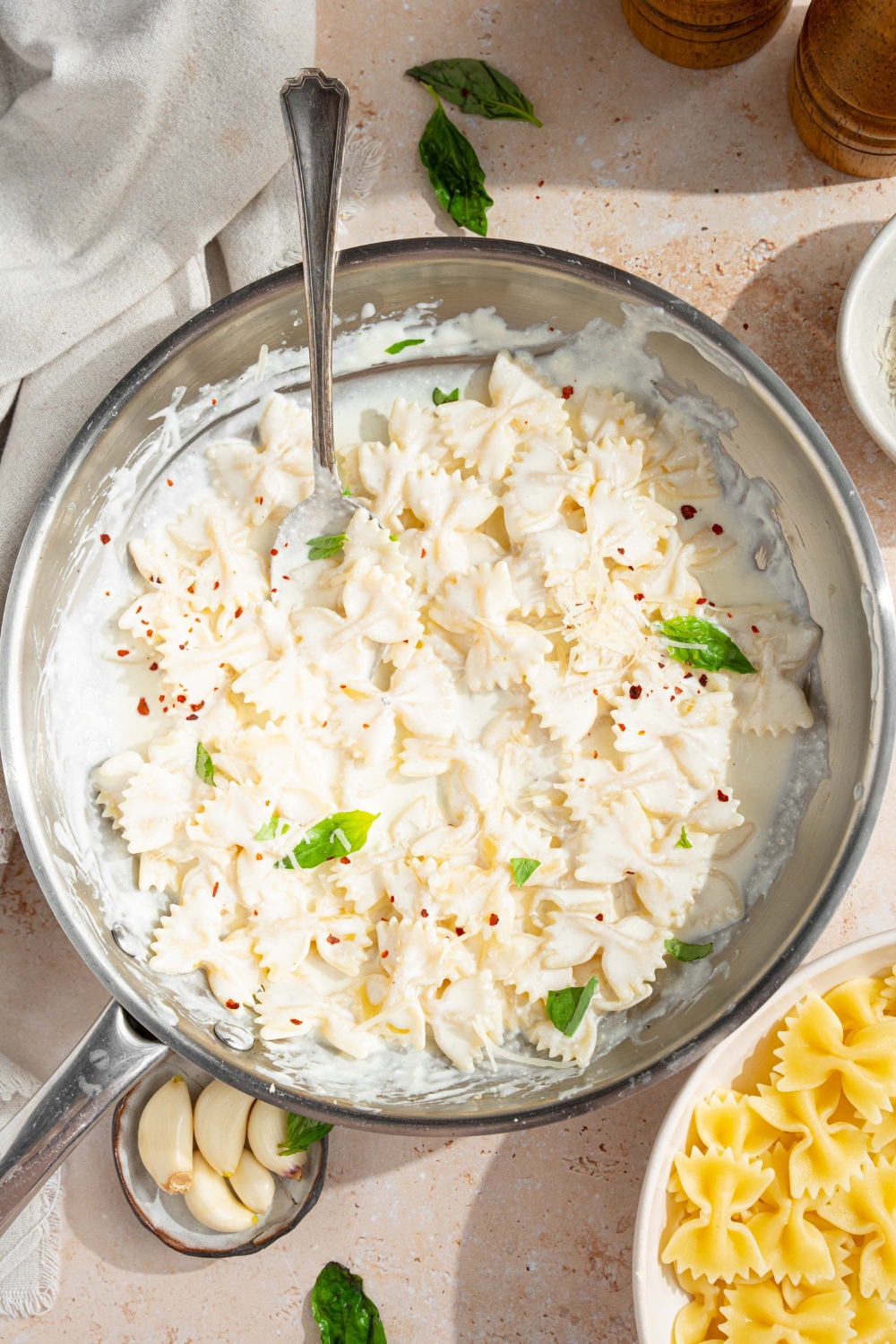 A skillet with bowtie pasta tossed in alfredo sauce. The pasta is garnished with fresh parsley and crushed red pepper. There is a fork in the pasta. The skillet is on a tan counter with a bowl of garlic and bowl of bowtie pasta.
