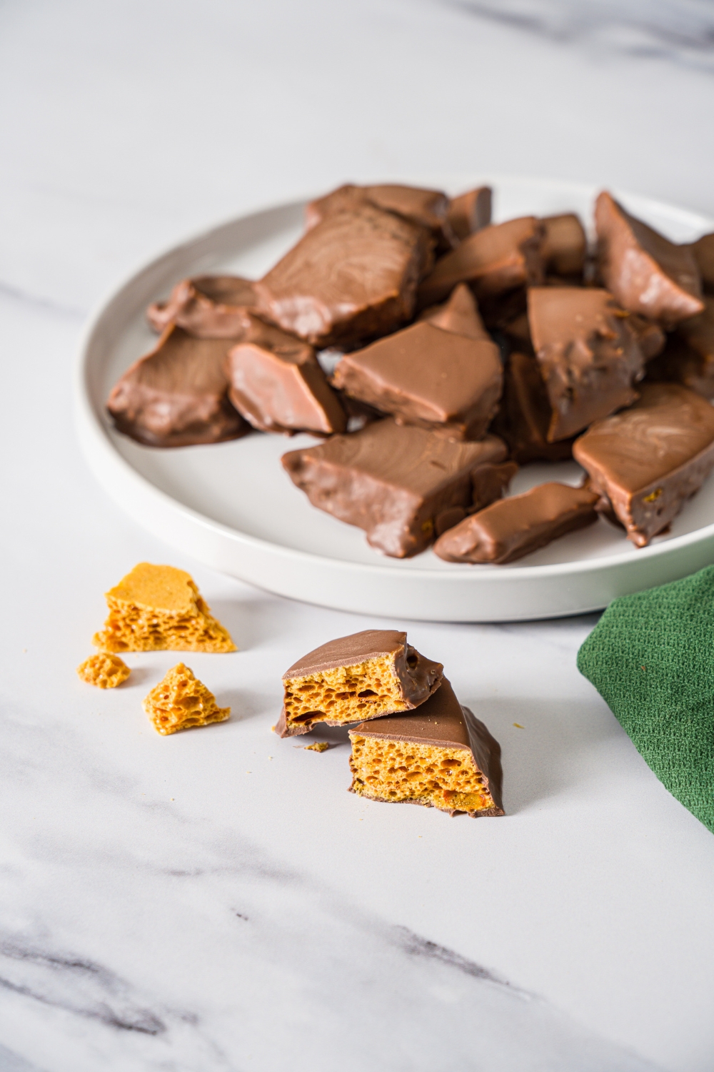 A white plate with homemade sponge candy on a marble counter. There is a piece of sponge candy sliced in half on the counter with a green cloth napkin.