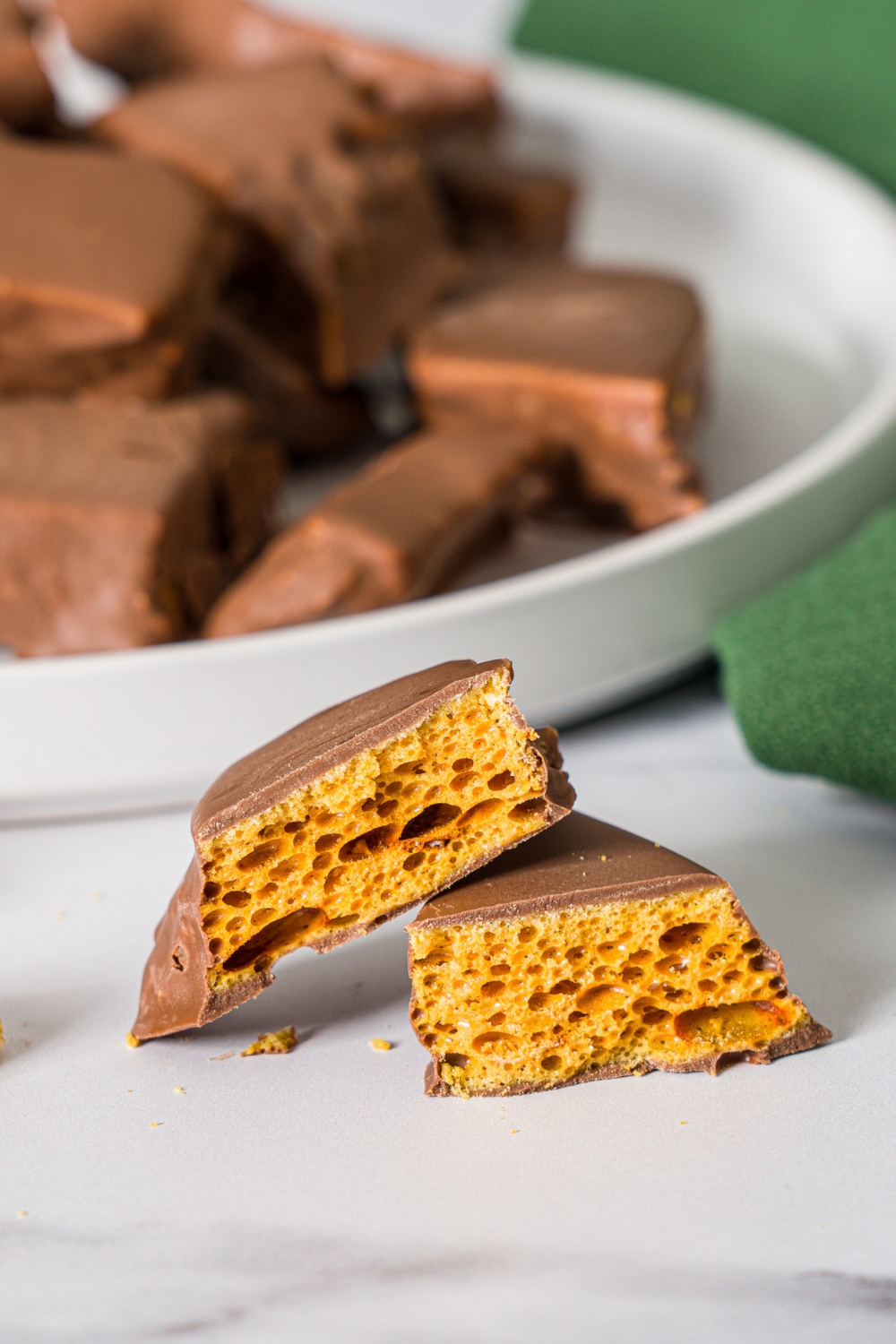 A sliced piece of sponge candy on a marble counter with a white plate of sponge candy and green cloth napkin.