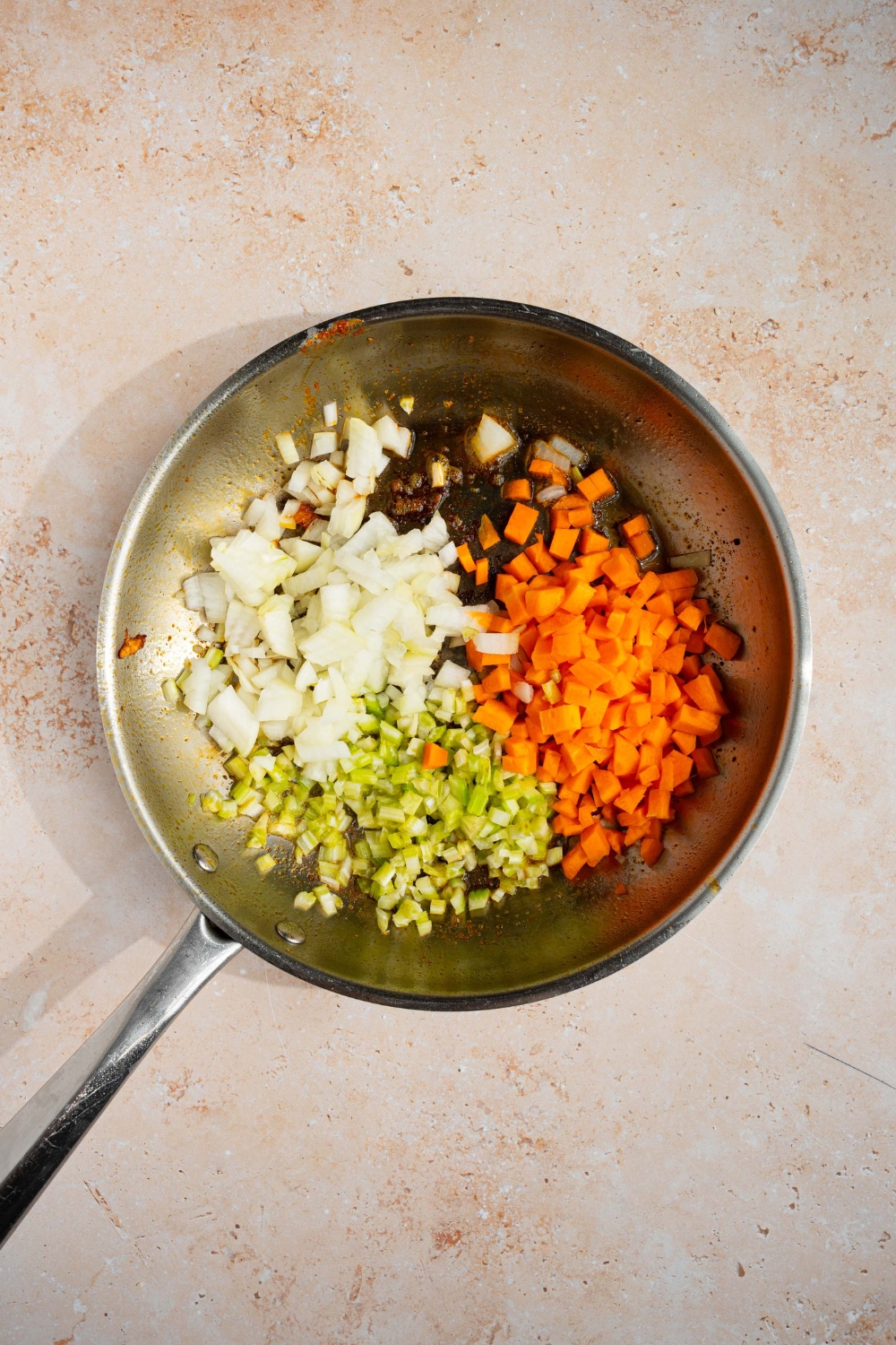A skillet with diced celery, onions, and carrots cooking in oil. The skillet is on a tan counter.