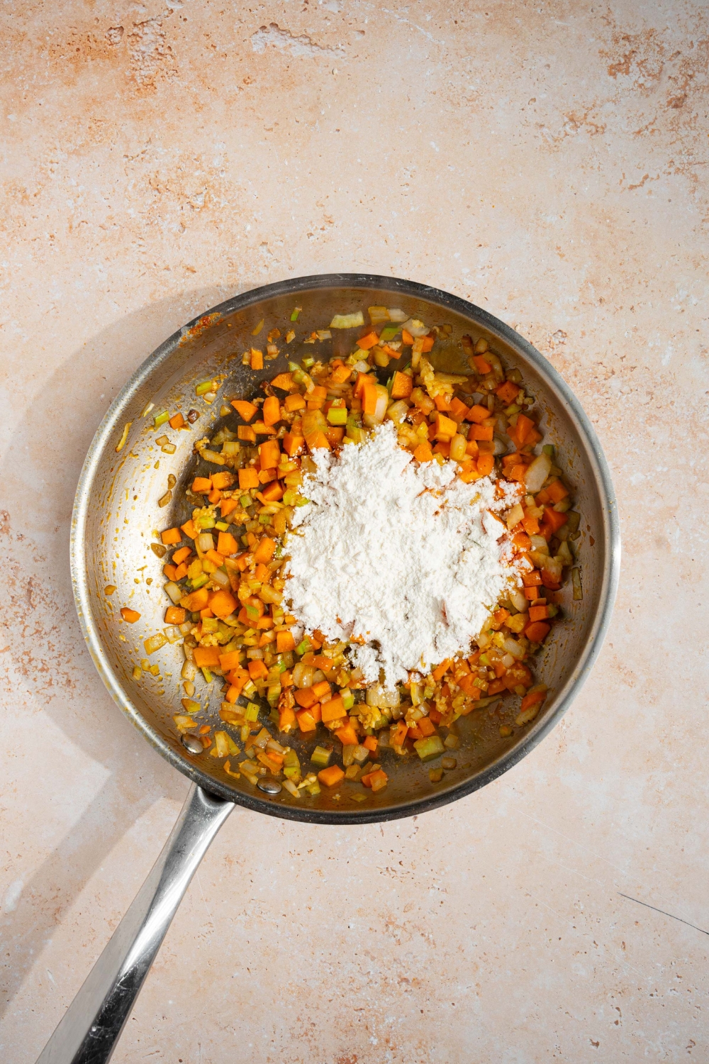 A skillet with diced vegetables cooked in oil and topped with flour. The skillet is on a tan counter.