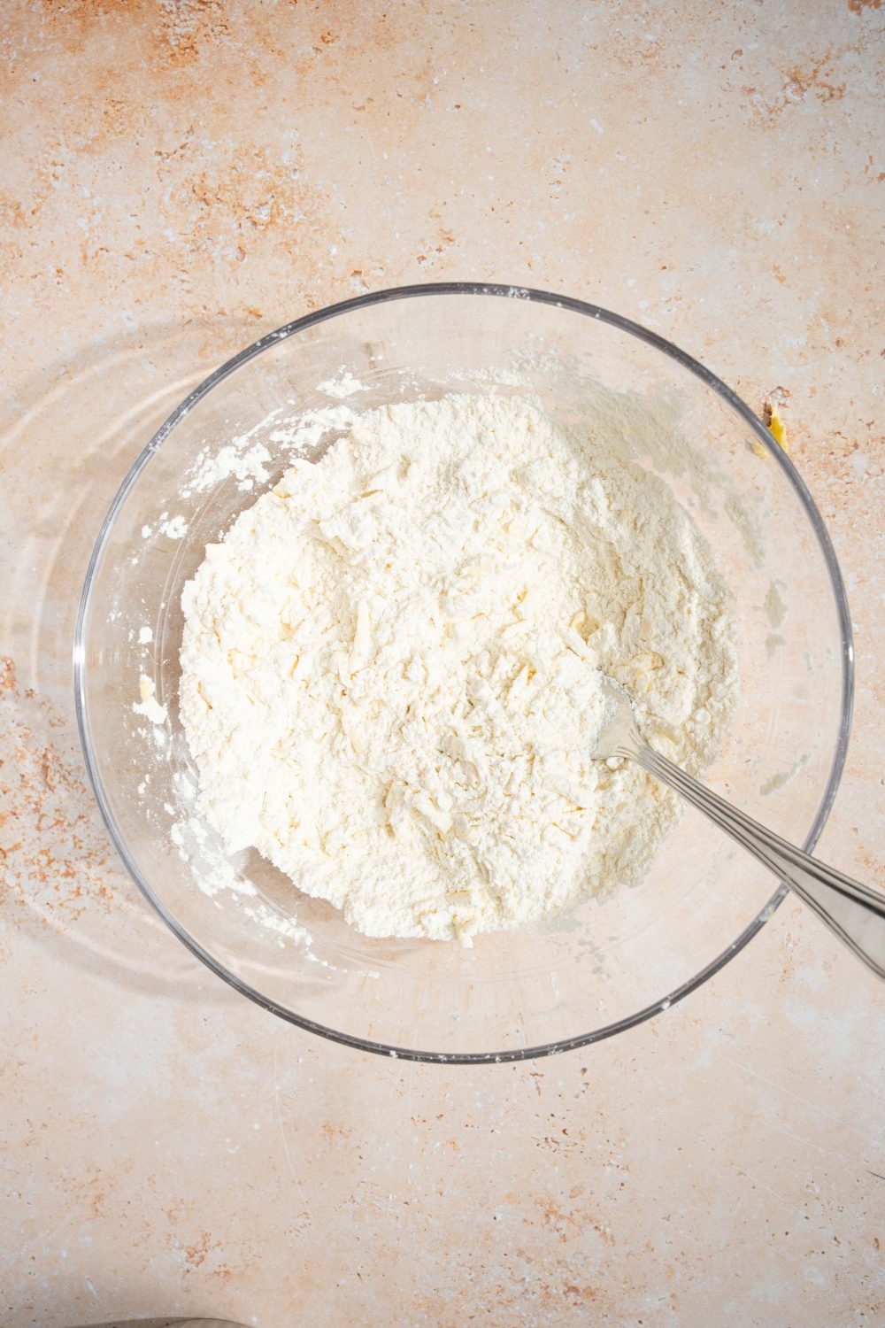 A glass bowl with a spoon mixing ingredients to make cheddar bay biscuit dough including flour, sugar, and grated butter. The bowl is on a tan counter.